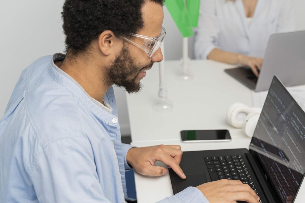 A person wearing glasses is typing on a laptop displaying lines of code at a white desk. Another person is using a laptop in the background. A phone, headphones, and papers are also on the desk.