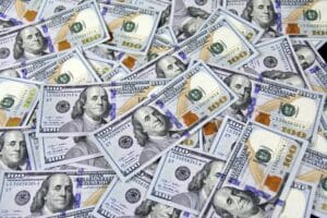 A large pile of U.S. one hundred dollar bills is spread out, showing multiple images of Benjamin Franklin and various security features on the banknotes.