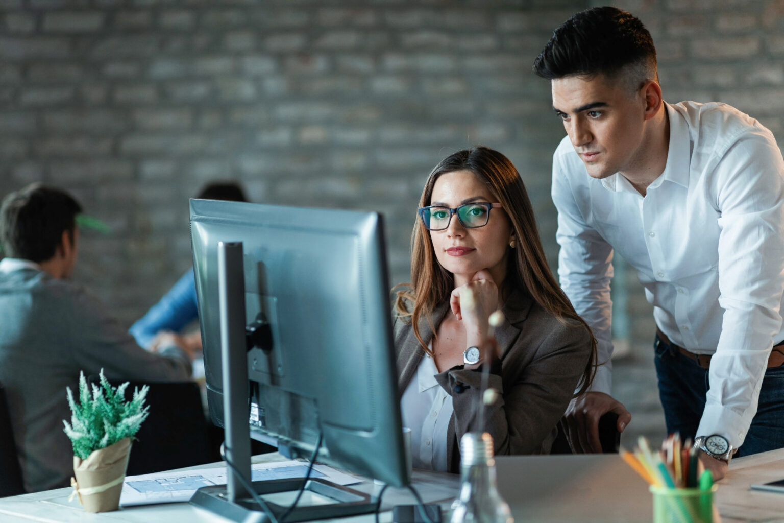 Two people in business attire look intently at a computer monitor in an office setting. The woman is seated with her hand on her chin, while the man stands beside her, leaning forward. Other workers are visible in the background.