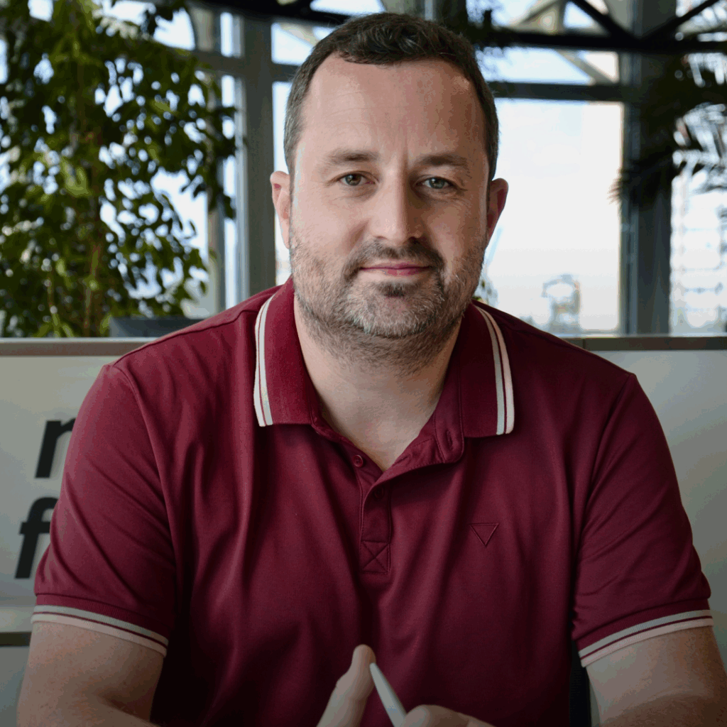A man with short dark hair and a beard, wearing a maroon polo shirt, sits indoors in front of a glass wall and plants, looking at the camera with a neutral expression.