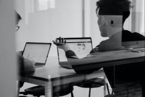 Two people sit at a table with open laptops in front of them, engaged in discussion. The image is black and white, and their reflections are visible in the glass partition. Office equipment is visible in the background.
